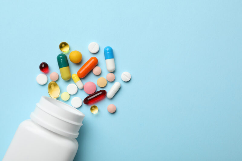Tabletten und Kapseln aus einer Medikamentendose auf hellblauem Grund